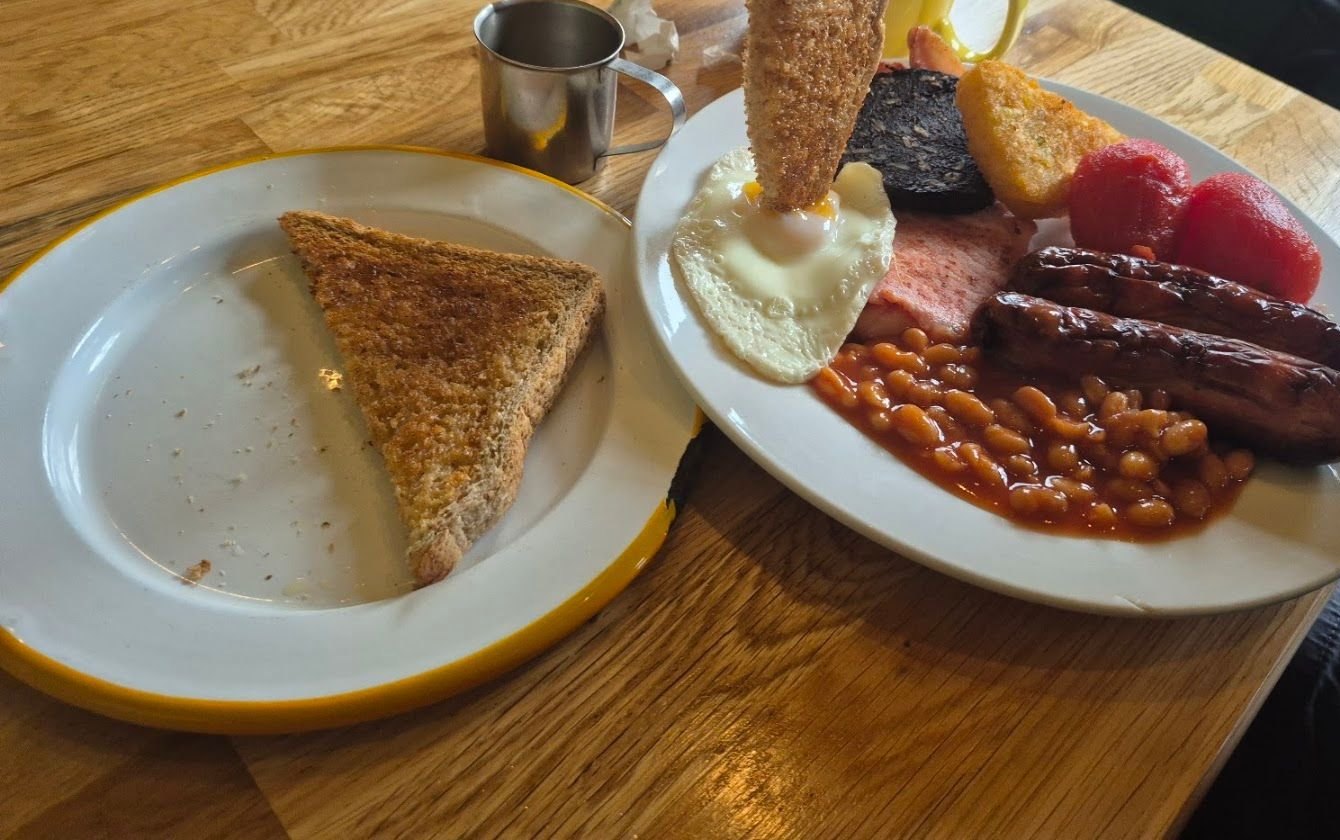 A cooked breakfast on a white plate with a yellow rim: sausages, bacon, black pudding, a fried egg, beans and tomatoes, with toast on a second plate