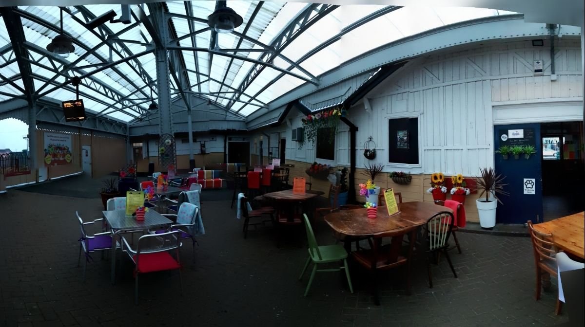 Café tables and brightly coloured chairs under the glass-roofed station concourse, with timber-boarded walls